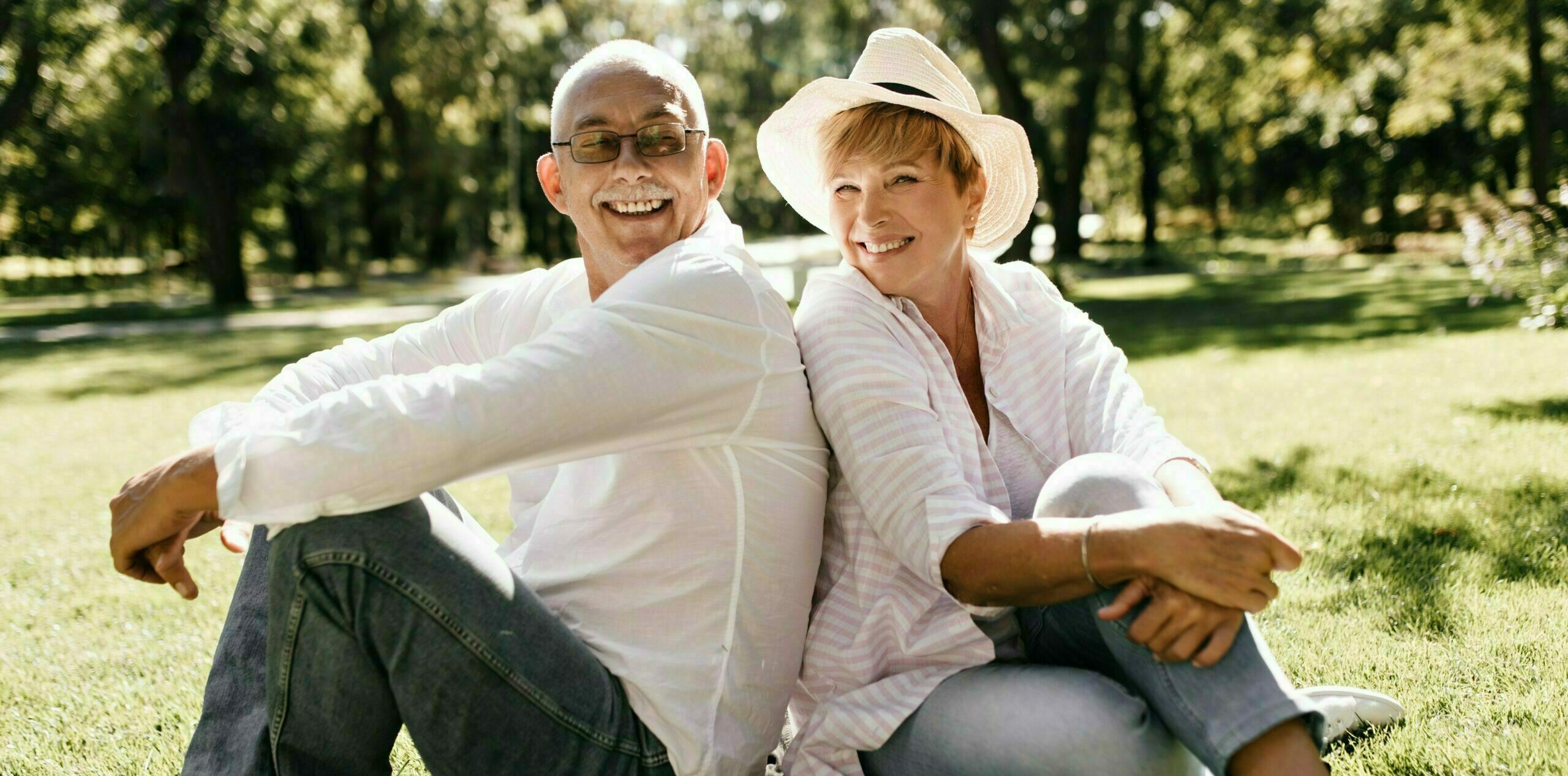 Positive lady with cool hat in striped stylish blouse and jeans smiling and sitting on grass with man in eyeglasses and light shirt outdoor..